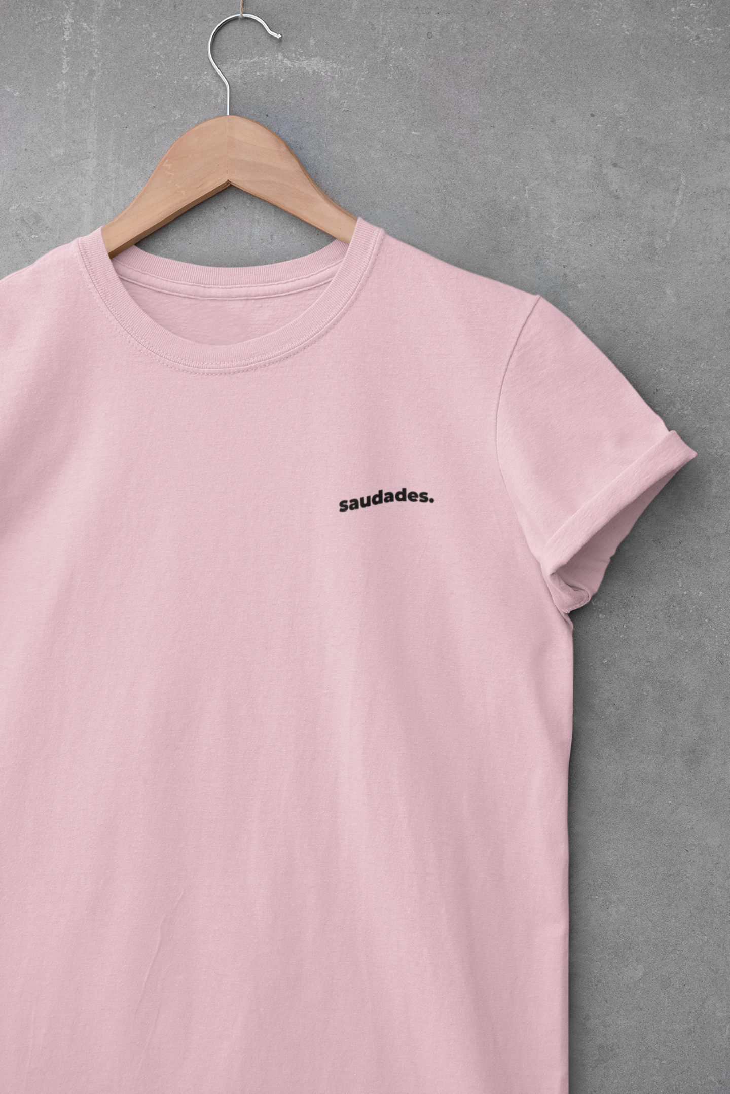 Pink t-shirt with 'saudades.' text on a hanger against a gray background