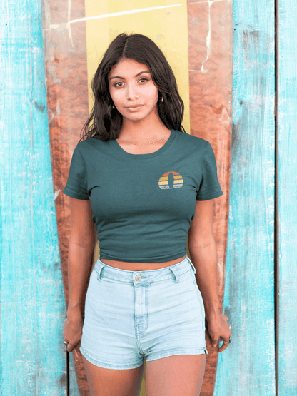 Woman wearing a fitted teal Nazare Surf Minimal Organic T-shirt with a vintage surf logo, posing in front of a surfboard. Sustainable and stylish beachwear.