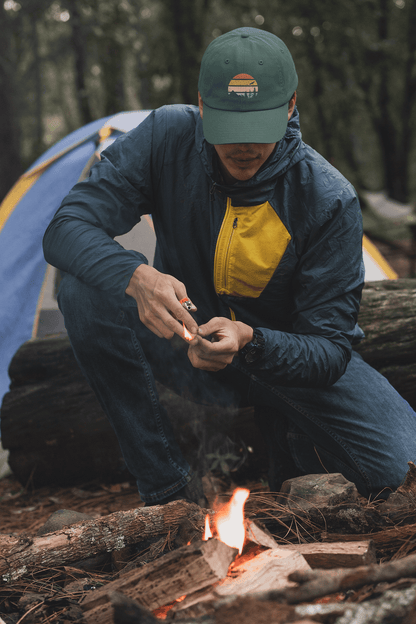 Madeira Vintage Cap in green with a mountain logo, worn by a camper lighting a fire. Perfect for outdoor adventures and rugged exploration.