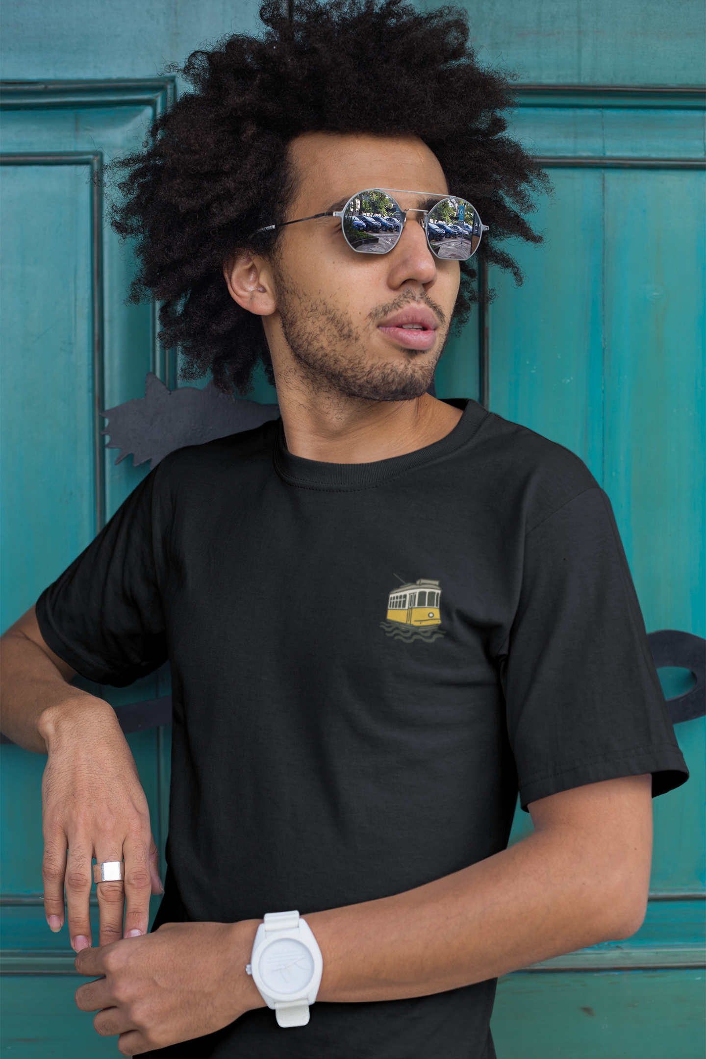 Man wearing a black t-shirt with a small graphic design, sunglasses, and a white watch against a teal door.